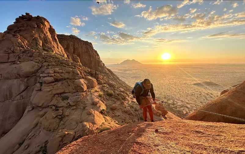 Climber on a rope high on Spitzkoppe, with the sun low over the plains