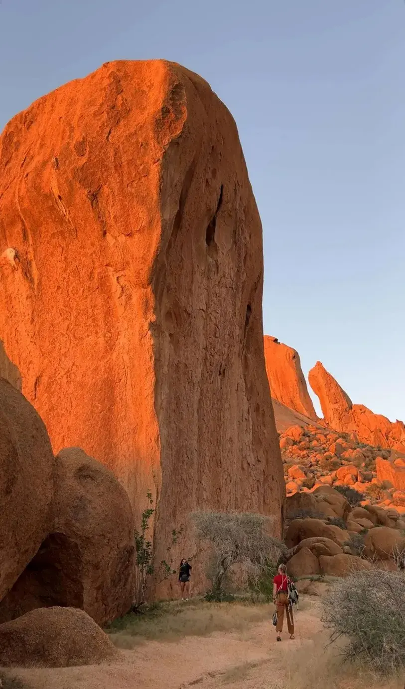 Two walkers at the foot of a tall granite tower at Spitzkoppe, lit orange by the evening sun